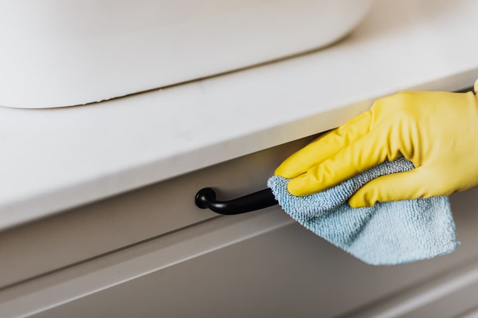 A gloved hand wiping a cabinet handle with a blue microfibre cloth